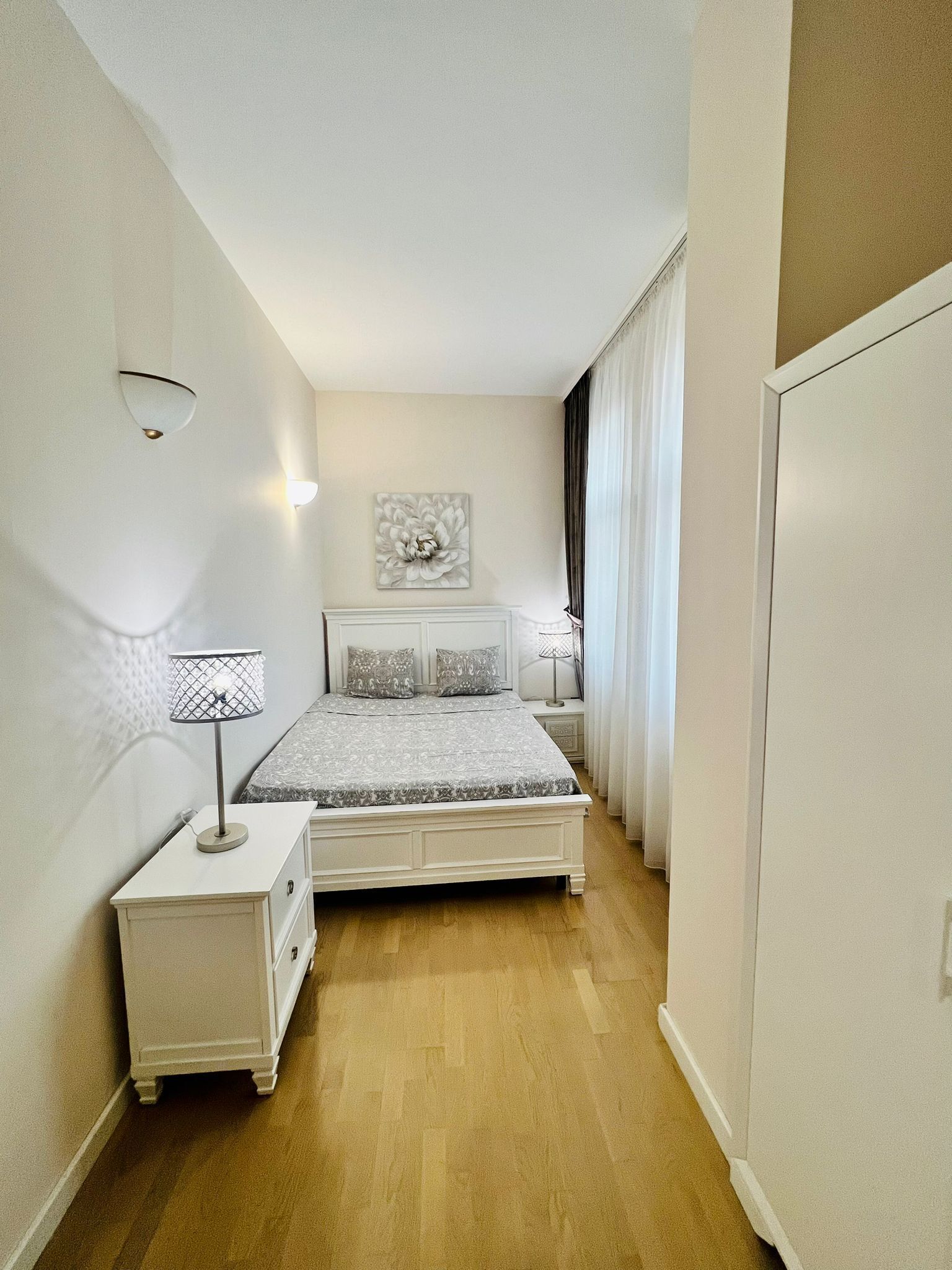 A narrow, well-lit bedroom with a bed, bedside lamps, and a wall-mounted artwork.