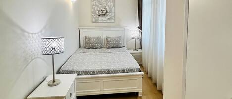 A narrow, well-lit bedroom with a bed, bedside lamps, and a wall-mounted artwork.