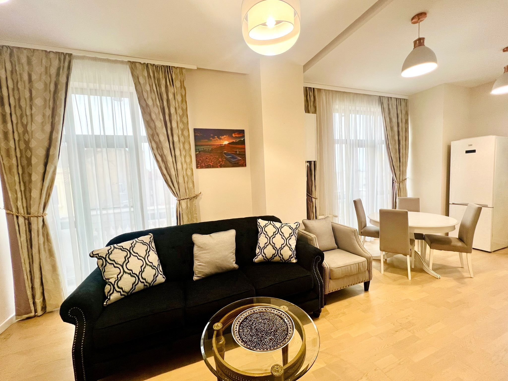 Una sala de estar moderna con un sofá negro, cojines estampados, una mesa de centro de vidrio y un área de comedor con una mesa y sillas blancas.
