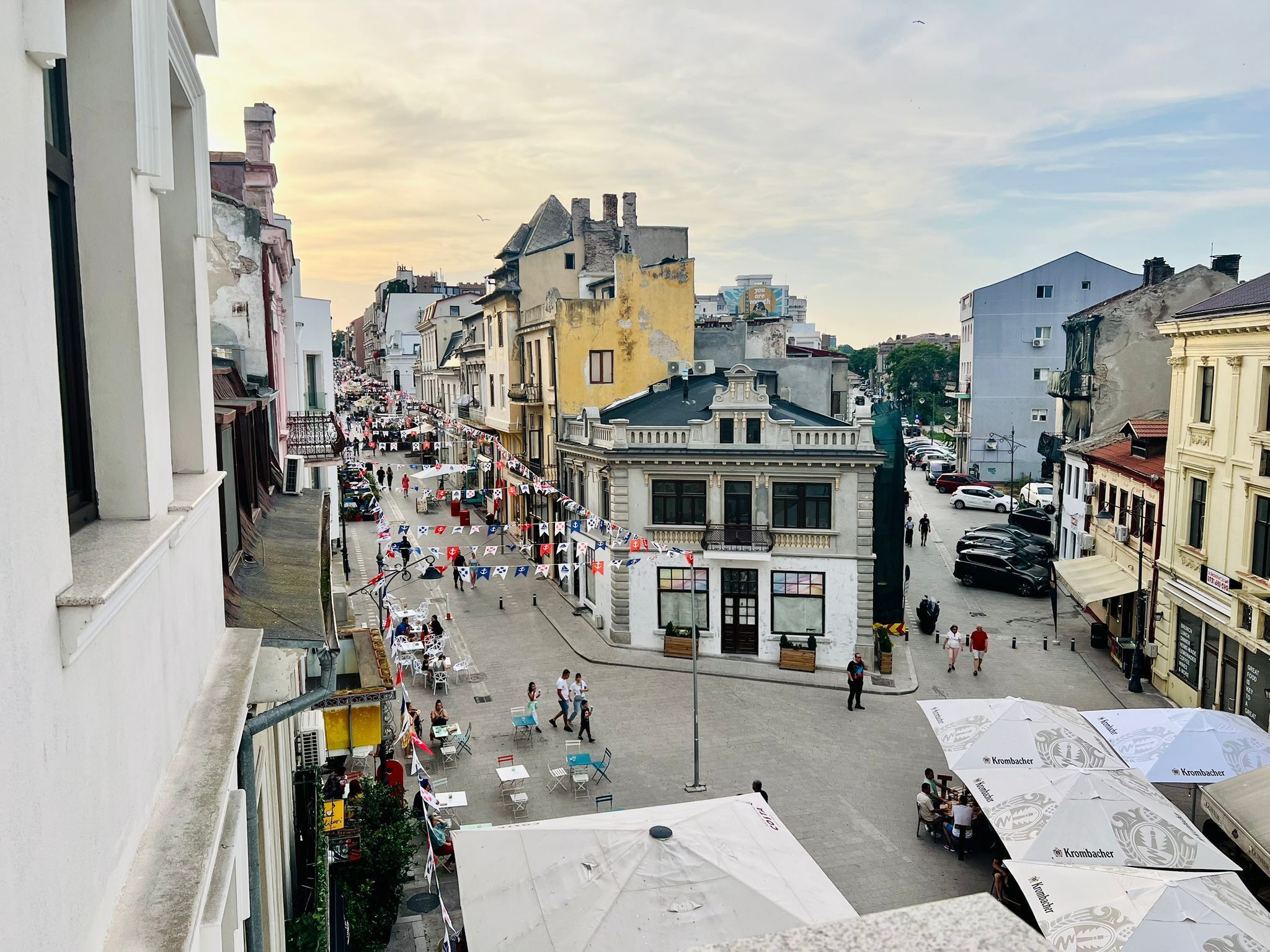 Rua com edifícios, mesas ao ar livre e bandeiras.