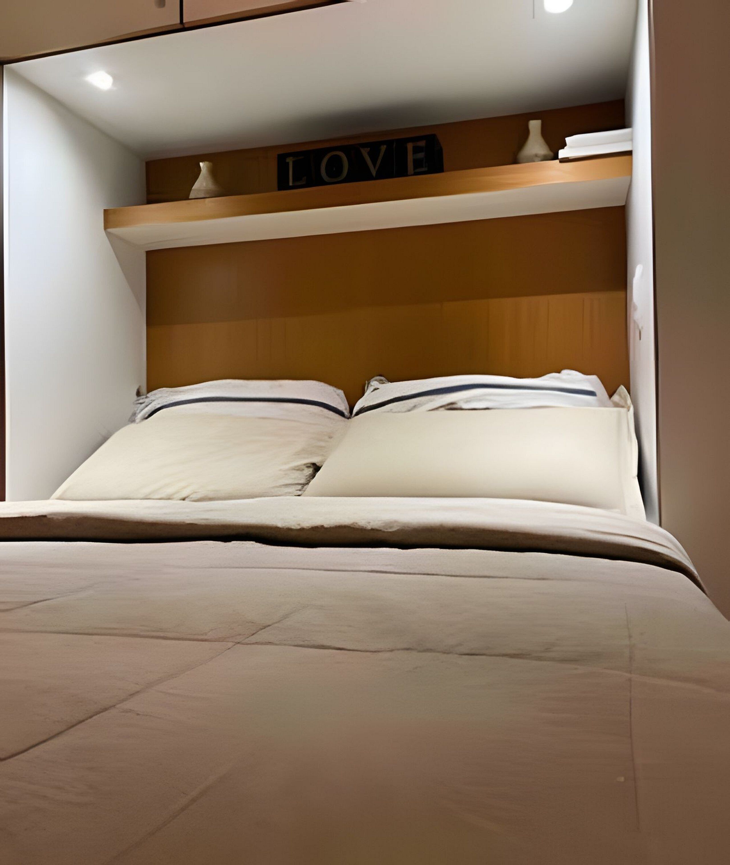 Uma cama com cabeceira de madeira, uma prateleira acima com a placa 'LOVE' e um vaso branco na prateleira.