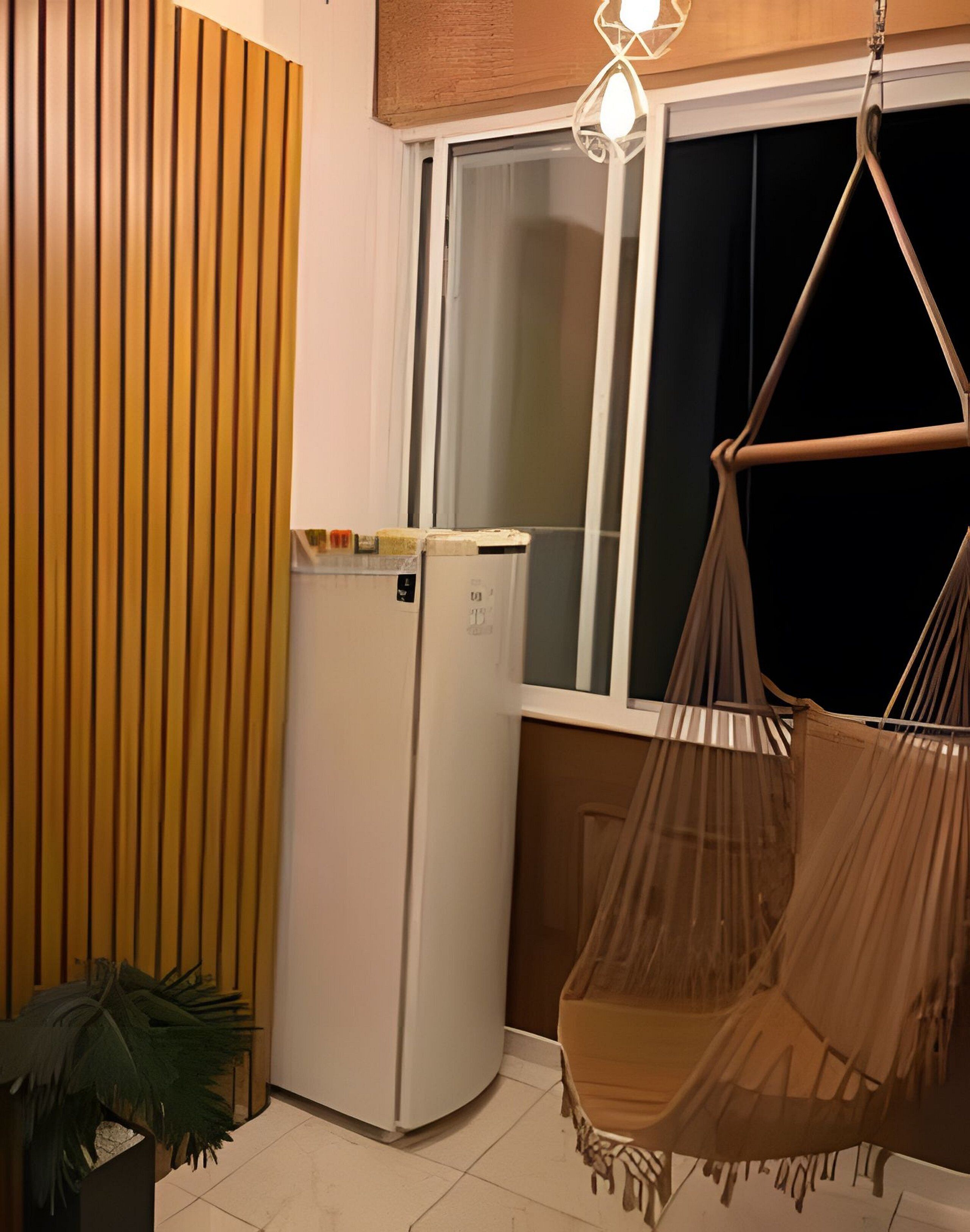 A compact room with a white refrigerator, a hanging chair, and a window with blinds.