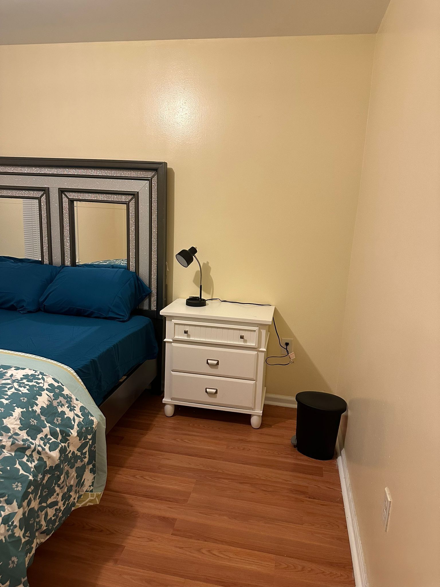 Un dormitorio con cama, mesita de noche, lámpara y cubo de basura.