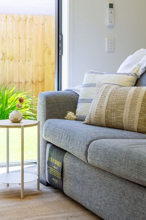 A grey sofa with patterned cushions, a small white side table with a vase, and a view of a wooden fence outside.