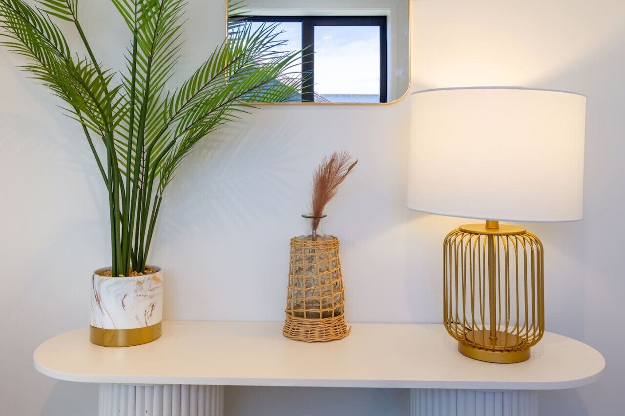 Une console blanche ornée d’une plante en pot, d’un vase en osier contenant des herbes de la pampa séchées et d’une lampe de table à abat-jour doré.