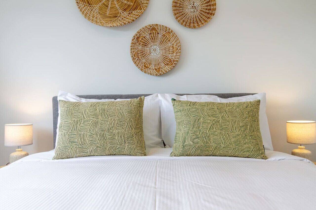 Una cama bien hecha con ropa blanca y dos almohadas verdes con diseño de hojas. Encima de la cama, tres adornos de pared tejidos.