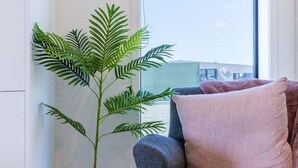 Une pièce avec un canapé gris, une plante en pot posée sur un tabouret en bois et une fenêtre donnant sur des immeubles.
