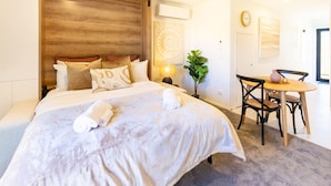 Habitación de hotel con una cama grande, una mesita de noche con lámpara, una pequeña mesa con sillas y una planta.