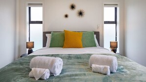 A neatly made bed with green and white bedding, two pillows, and two towels on the bed. There are two bedside lamps on either side of the bed.