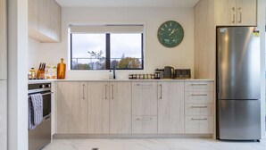 A modern kitchen with light wood cabinets, a stainless steel refrigerator, a clock, and a window with a view.