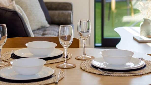 Una mesa de comedor dispuesta con platos, tazones y copas de vino, con vistas a un jardín a través de un amplio ventanal.