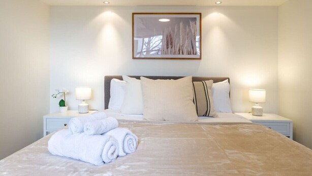 Una cama bien hecha con toallas blancas enrolladas, un cuadro grande enmarcado encima y dos lámparas de noche en mesitas blancas.