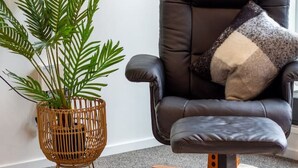 Una sala moderna con un sillón de cuero negro, un atril tejido para plantas y un cojín gris.