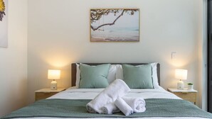 Una cama bien hecha con una colcha verde, dos almohadas y una toalla enrollada. Hay dos mesitas de noche con lámparas y un cuadro colgado en la pared que representa una playa.