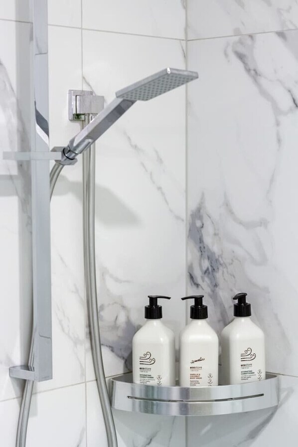 A bathroom with a shower, a glass shelf holding three bottles, and a marble wall.