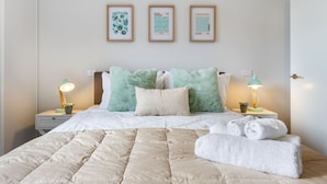 Uma cama bem arrumada com um edredom bege, duas almofadas verdes e uma almofada branca. Há três quadros na parede acima da cama.
