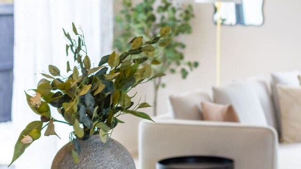 A modern living room with a grey textured vase on a wooden coaster, a round table, and a white sofa with cushions.