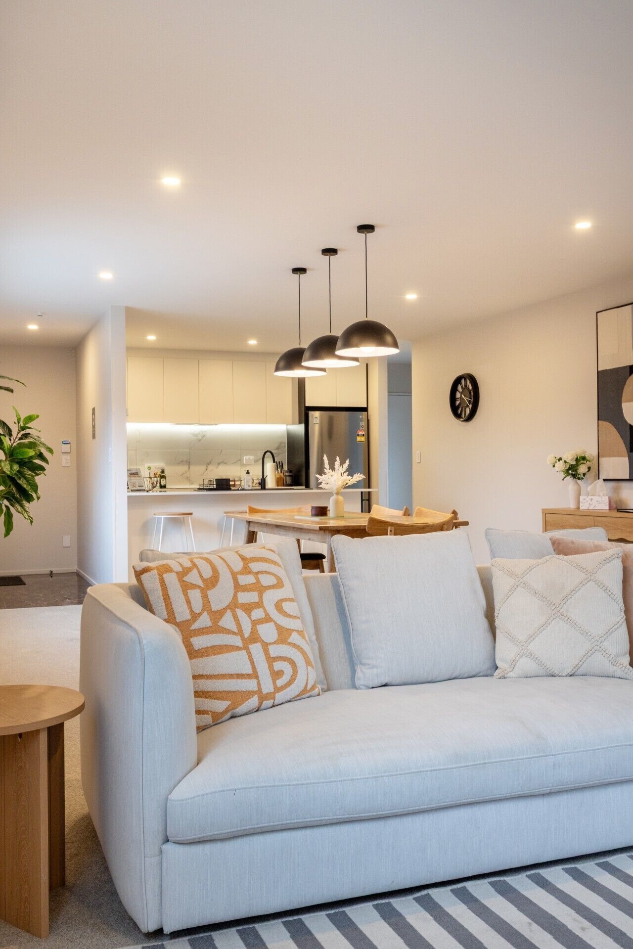 Una sala moderna con un sofá, una mesa de comedor, lámparas colgantes, una cocina al fondo y un reloj en la pared.