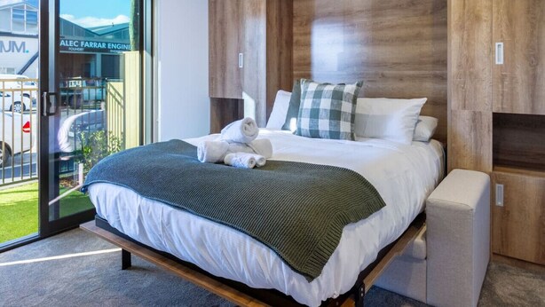 A modern bedroom with a large bed, a bench at the foot, and a view of a building outside. - Single Level Studio Escape on Wilmer St - Central (Christchurch)