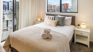 Habitación de hotel con una cama grande, ventana con vista a la ciudad, balcón con una silla y lámparas de noche.