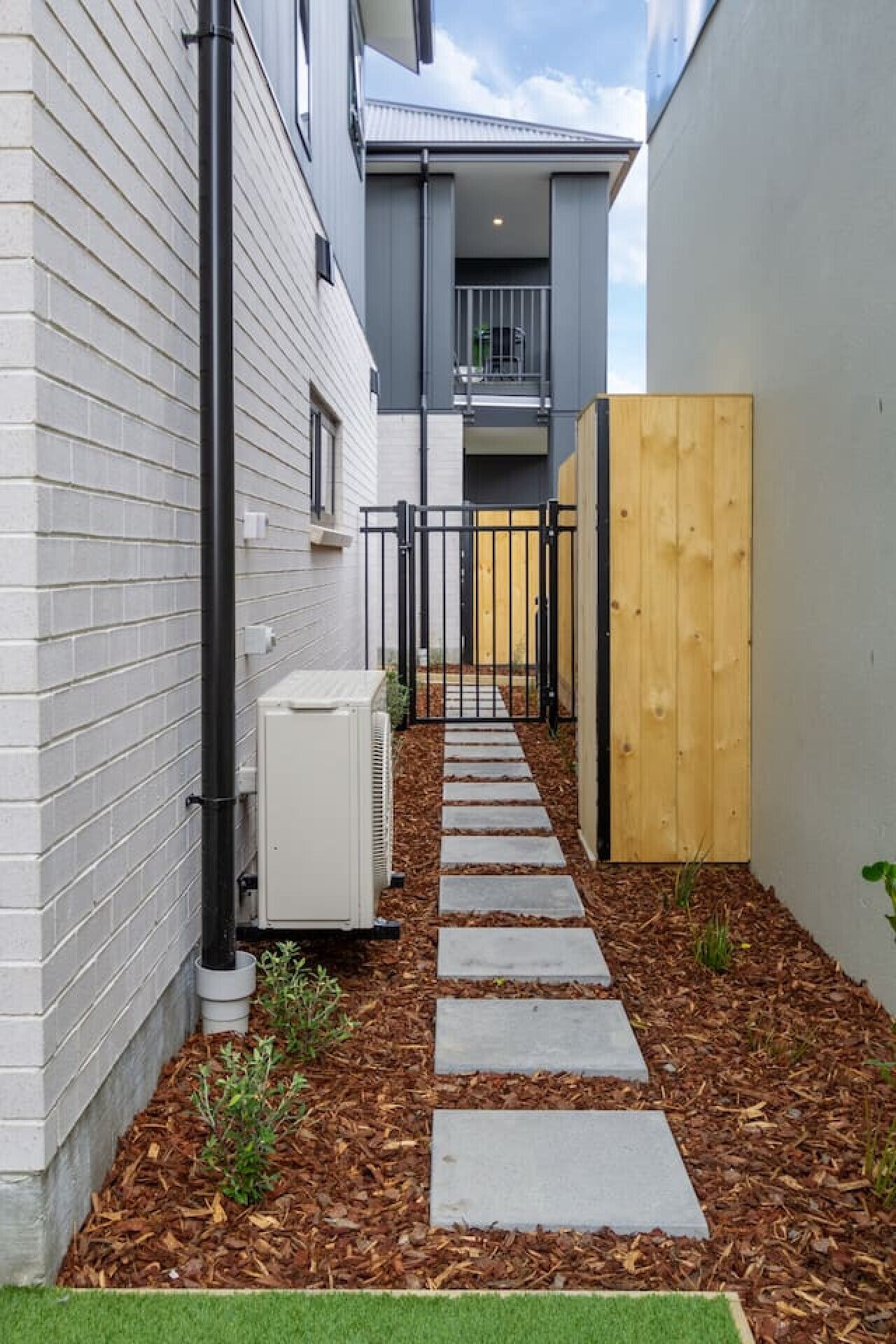 Un sendero estrecho que conduce a una entrada con portón, flanqueado por un edificio de ladrillo blanco y una estructura gris con balcón.