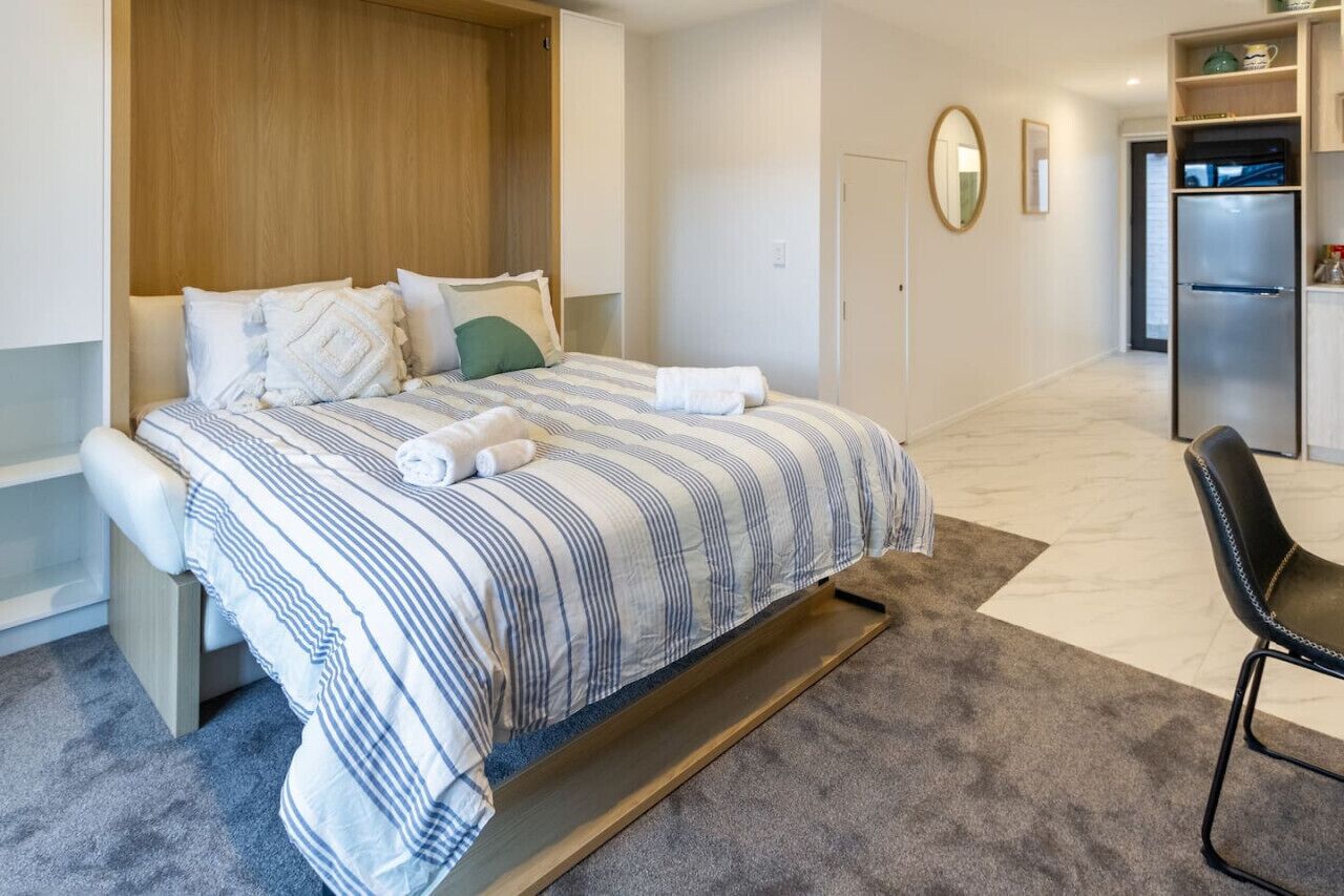 A bedroom with a bed, a wooden headboard, a small desk, a chair, a refrigerator, and a bathroom door.