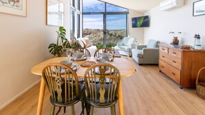 A dining area with a wooden table set for four, surrounded by chairs. A large window offers a view of the outdoors. - The Keep - Waimairi Beach (Christchurch)