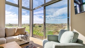 A modern living room with large windows, a sofa, a chair, and a flat-screen TV. - The Keep - Waimairi Beach (Christchurch)