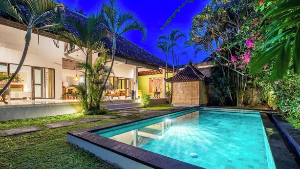 A swimming pool surrounded by a well-maintained lawn, a patio area with outdoor seating, and a traditional-style building with a tiled roof. - Villa Amaro - 2 Bedrooms (Seminyak)