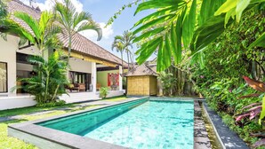 A swimming pool surrounded by lush greenery and a traditional house with a tiled roof. - Villa Amaro - 2 Bedrooms (Seminyak)