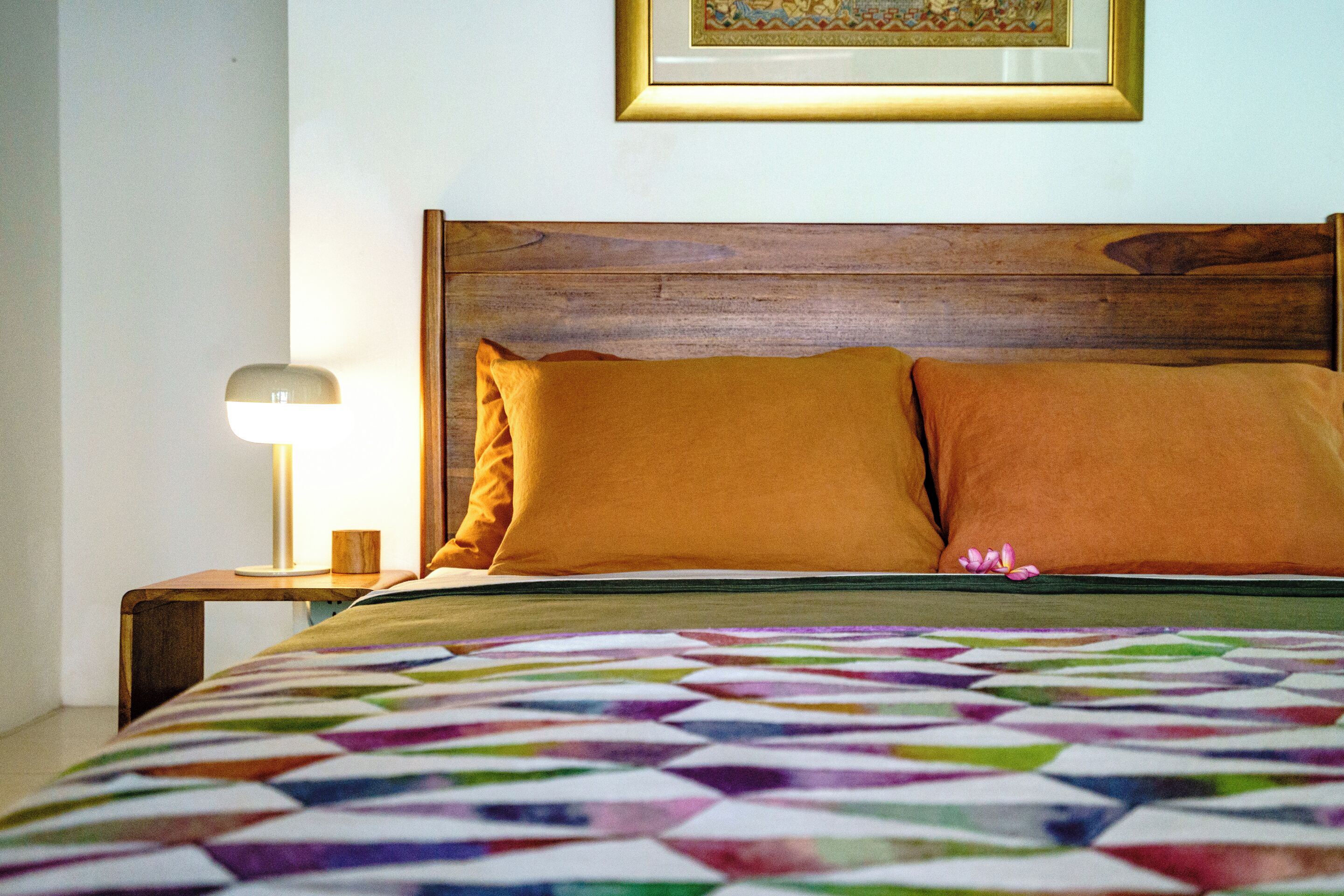 Una cama con cabecera de madera, dos almohadas anaranjadas, una colcha colorida y una lámpara de noche sobre una mesita de madera.