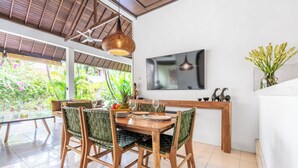 A dining area with a wooden table, green woven chairs, a hanging light fixture, and a TV mounted on the wall. - Villa Amaro - 2 Bedrooms (Seminyak)