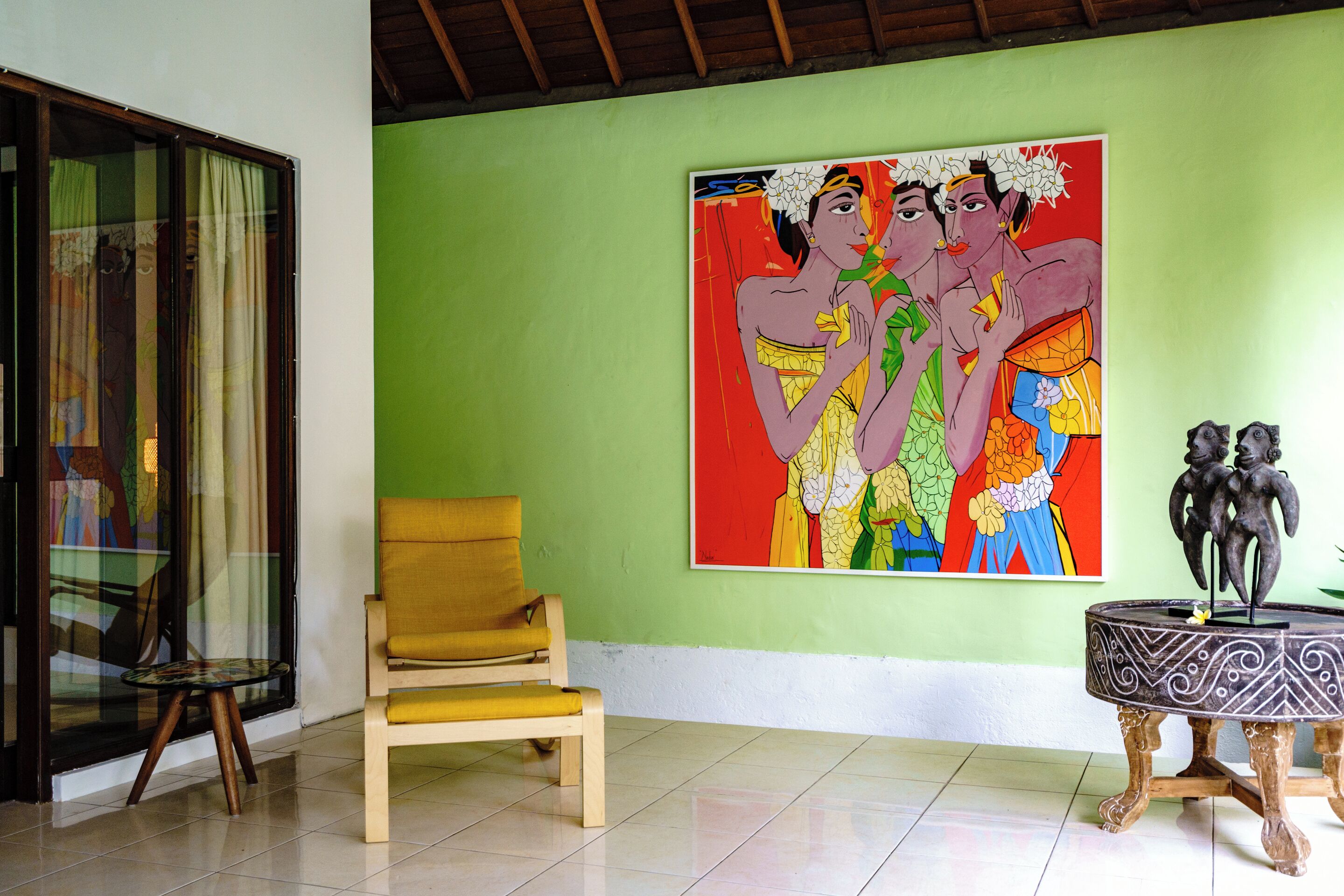 A room with a green wall, a wooden chair, a table with a decorative base, and a colorful painting.