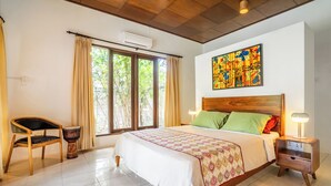 A bedroom with a bed, a chair, a drum, a nightstand, and a window with curtains. - Villa Amaro - 2 Bedrooms (Seminyak)