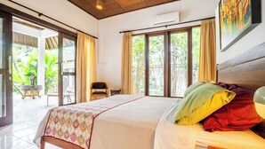 A spacious bedroom with a large bed, a wooden headboard, and a view of greenery through the windows. - Villa Amaro - 2 Bedrooms (Seminyak)