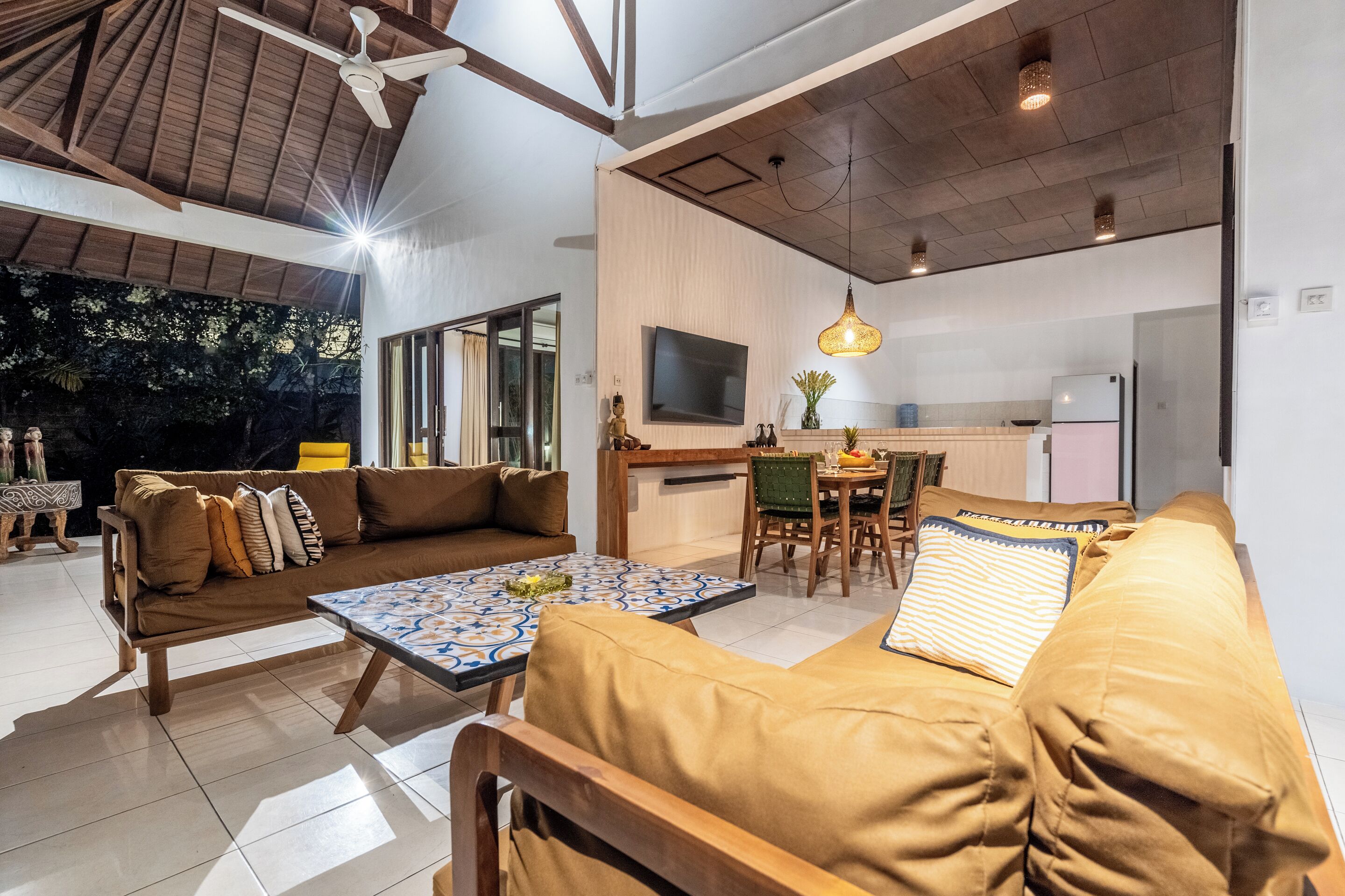 Ein geräumiger Wohnbereich mit Sofa, Couchtisch und Esstisch. Ein Fernseher ist an der Wand befestigt, ein Deckenventilator ist sichtbar.