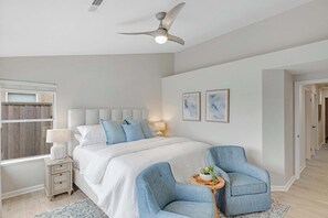Un dormitorio con una cama, dos sillas, una mesita de noche, un ventilador de techo y obras de arte en la pared.