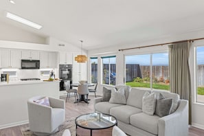 A modern kitchen with white cabinets, a dining area with a table and chairs, a gray sofa, and a large window with a view of the outdoors. - Stay Close to The Beach at Marina's Sand Piper! (Marina)