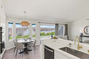A modern kitchen with a large island, a dining area with a round table and chairs, and large windows offering a view of a fenced yard. - Stay Close to The Beach at Marina's Sand Piper! (Marina)