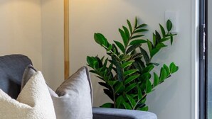 Un salon moderne avec un canapé gris, des coussins blancs et gris, une lampe sur pied, une plante verte et une fenêtre avec des rideaux blancs.