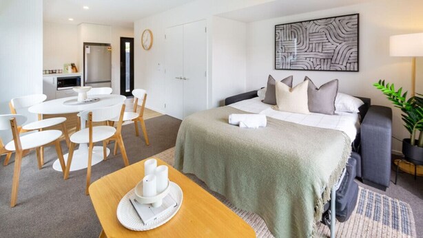 Une chambre d’hôtel moderne avec un lit, un coin repas comprenant une table et des chaises, une cuisine en arrière-plan et un grand tableau abstrait accroché au mur.