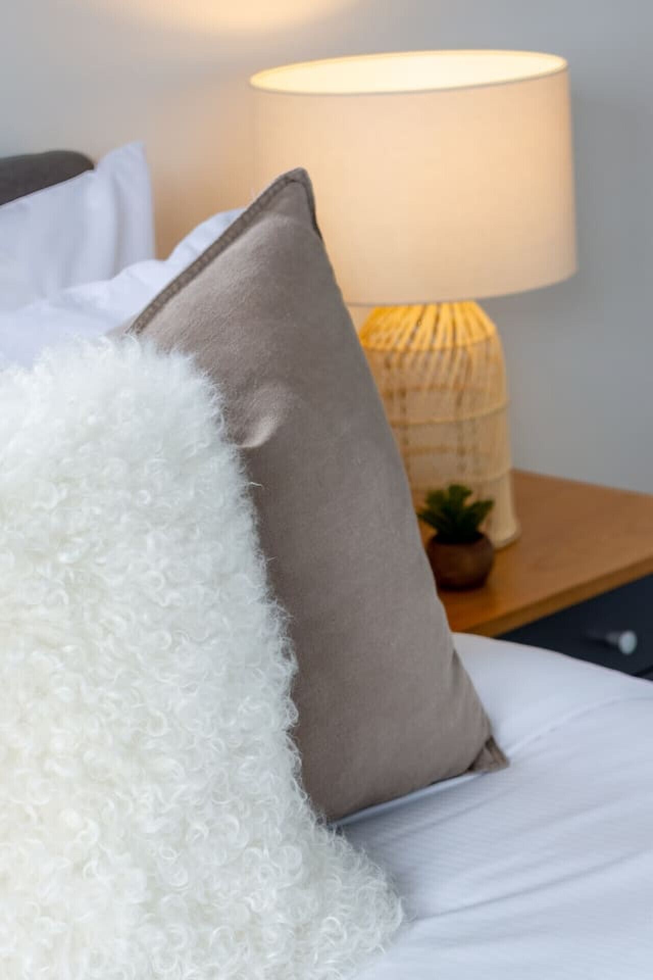 Una cama con un almohadón blanco y esponjoso y un almohadón beige, una lámpara de noche y una planta pequeña sobre una mesita de madera.