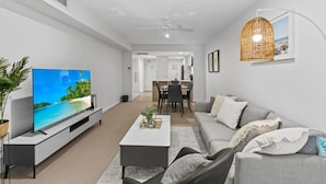 Een moderne woonkamer met een flatscreen televisie, een grijze bank, salontafel, en een eethoek op de achtergrond.