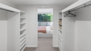 Een inloopkast met open planken en een kledingroede, die leidt naar een slaapkamer met een bed en uitzicht op groen buiten.