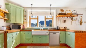 Une cuisine avec des armoires vertes, un évier blanc et un plan de travail en bois. Des luminaires sont suspendus au-dessus de l’évier, et une fenêtre offre une vue sur les bâtiments à l’extérieur.