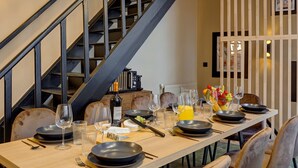 A dining area with a long table set for four, a staircase, and a kitchen area in the background. - TBS 242 - Exceptional triplex loft (Paris)
