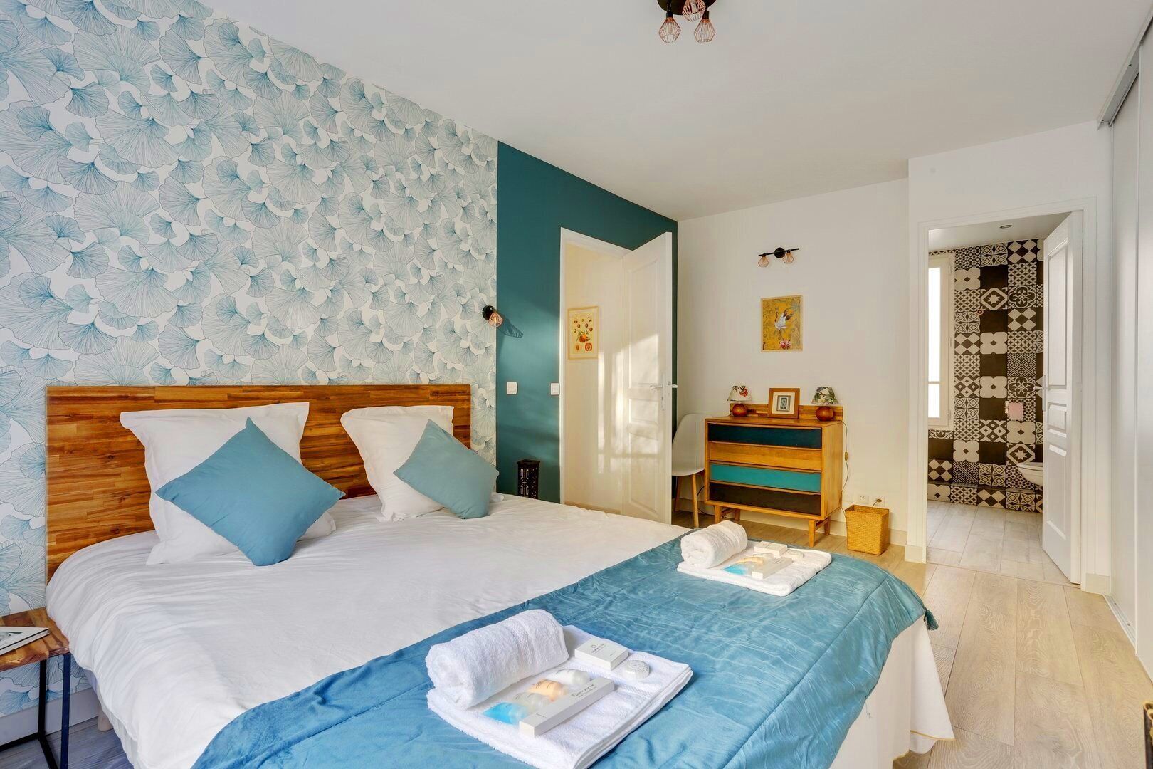 Un dormitorio con cabecera de madera, cama con ropa de cama blanca y azul, manta azul y una mesita de noche con lámpara.