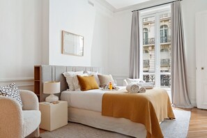 Et hotelværelse med en seng, et natbord, en lampe og udsigt til en bygning gennem vinduet.