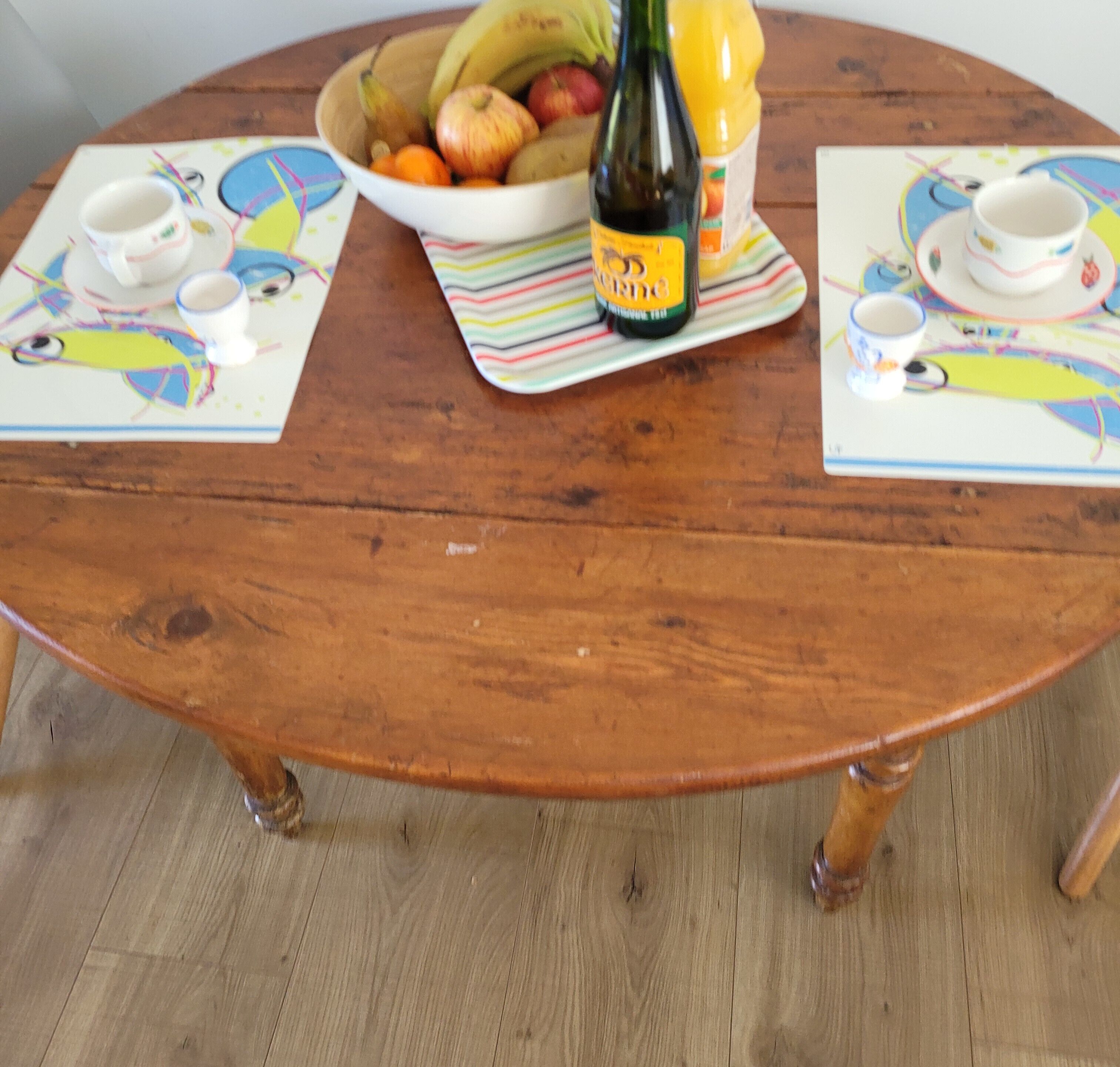 Una mesa redonda de madera con un frutero y una botella de vino encima.