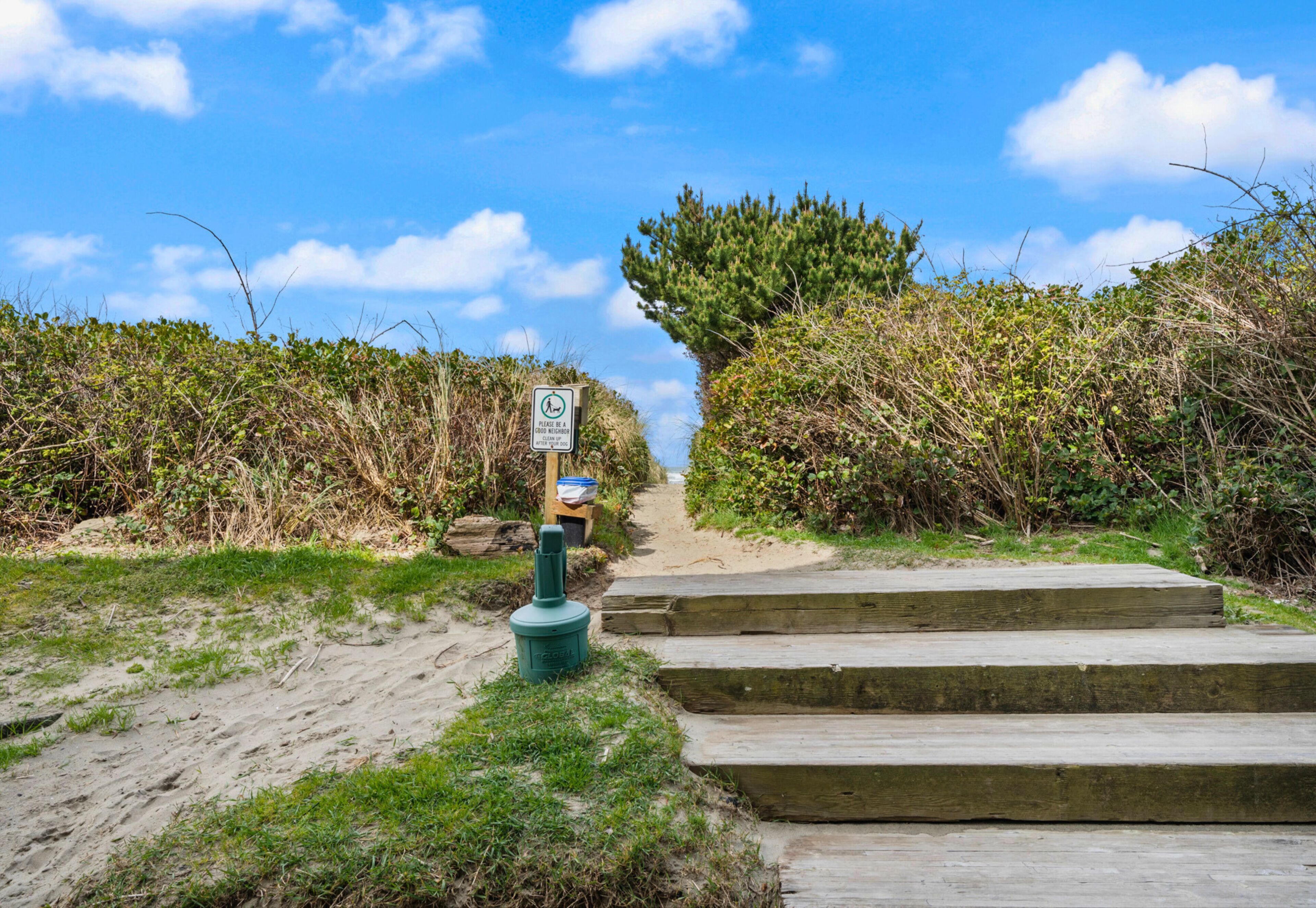 Un sentier sablonneux mène à un escalier en bois, et un panneau indique l’accès à la plage.